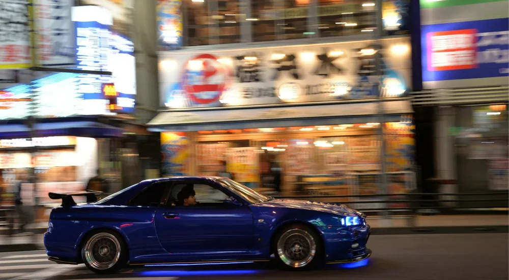 Modified blue Nissan Skyline GT-R cruising a neon-lit Tokyo street at night