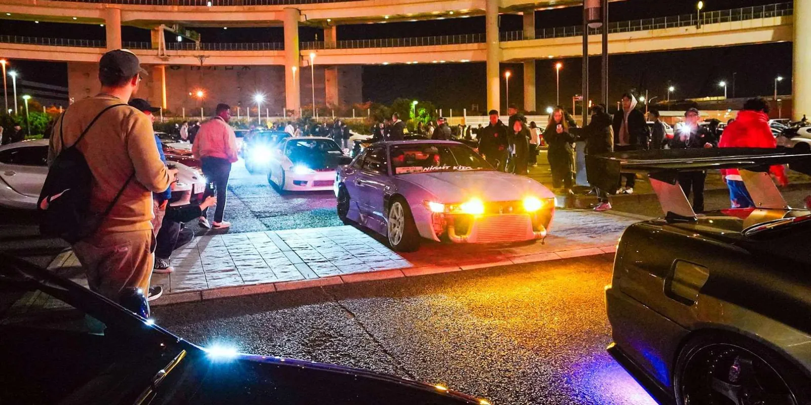 Lineup of modified Nissan Silvia S15s and tuner cars beneath Daikoku Parking Area's highway ramps