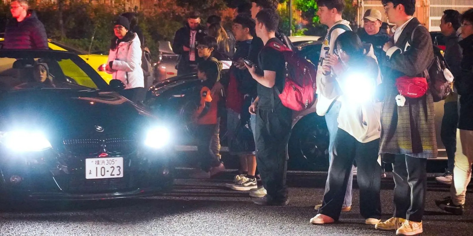 Car-meet crowd beside a modified BMW with its headlights on, gathered under the highway