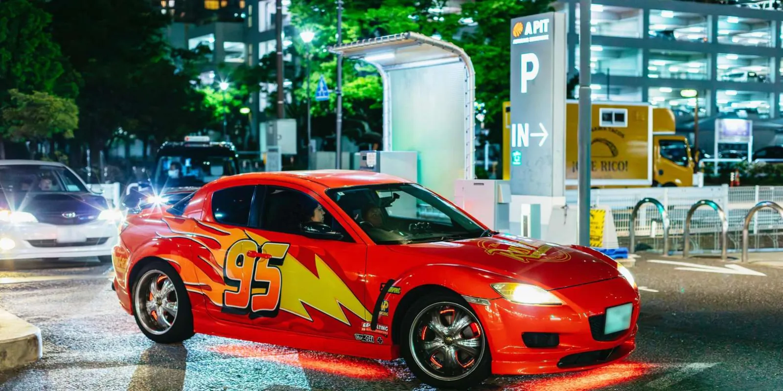 A right-hand-drive Mazda RX-8 in a racing livery pulling out of A-PIT Autobacs in Tokyo at night