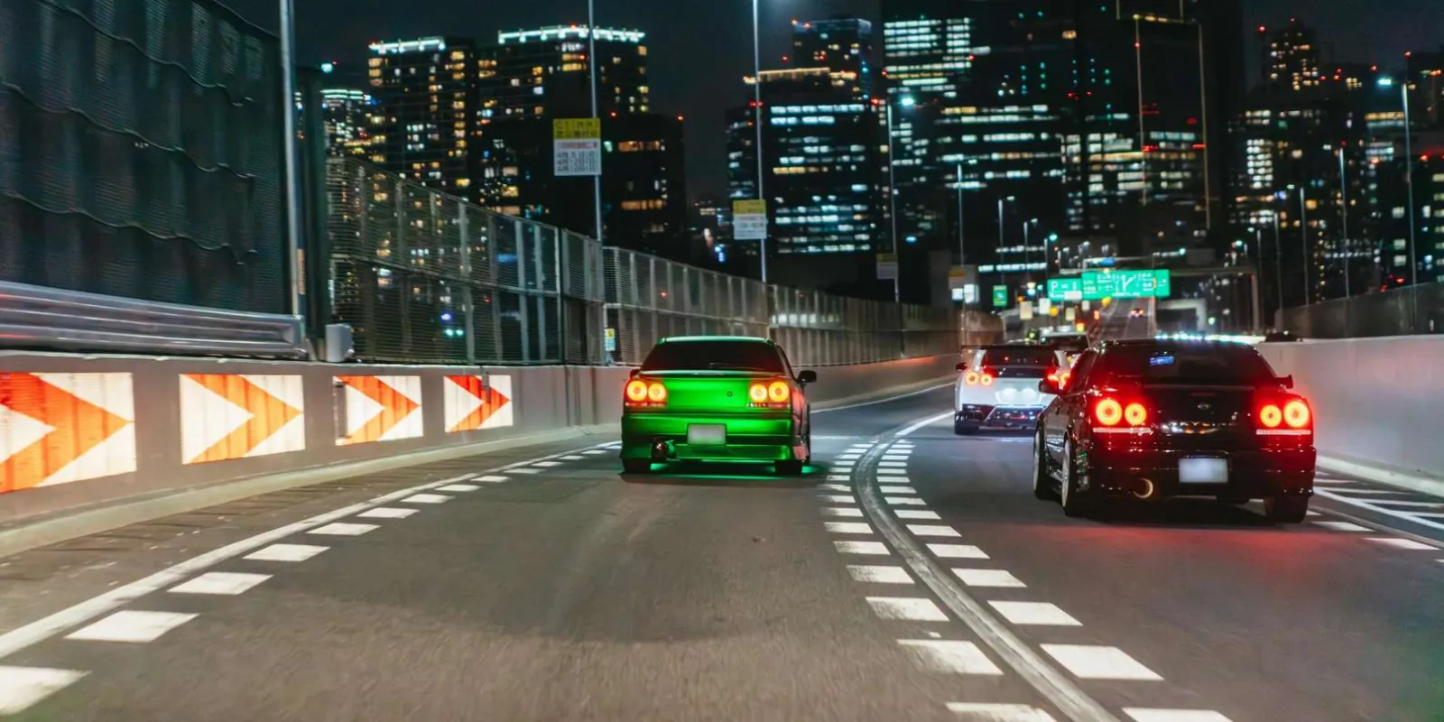 Tuned Nissan Skylines and a GT-R driving along a Tokyo expressway at night, with the city's towers ahead