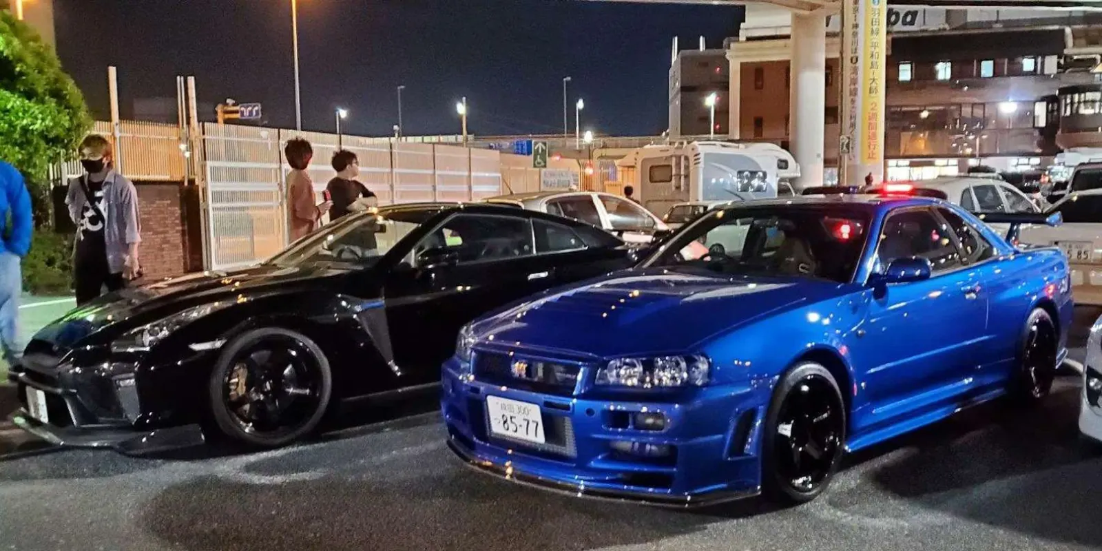 Modified Nissan Skyline GT-Rs and RX-7s parked along a row of illuminated garages at night